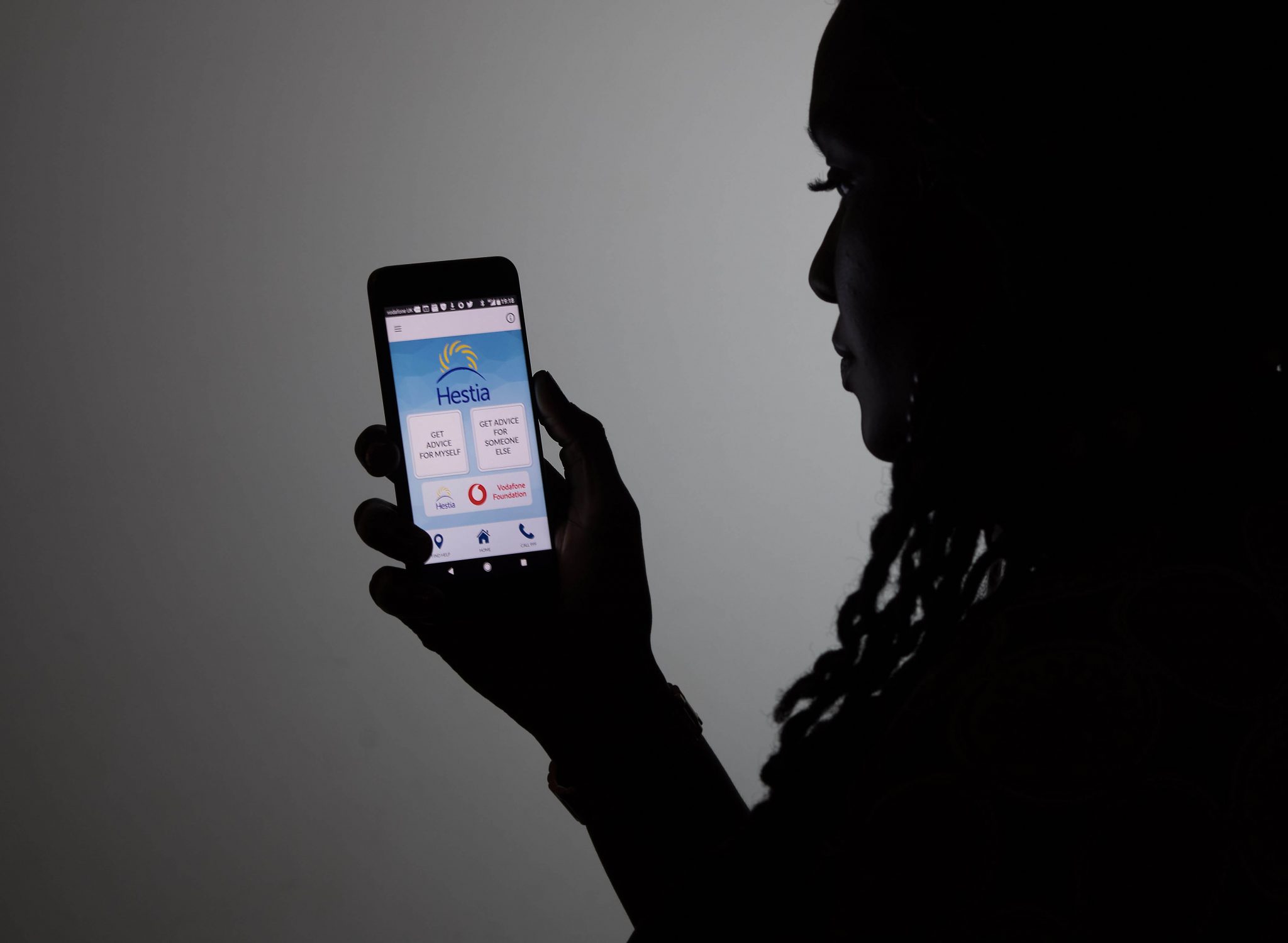 A woman holds up a smartphone showing the welcome screen for Bright Sky, an app created by the Vodafone Foundation in partnership with Hestia