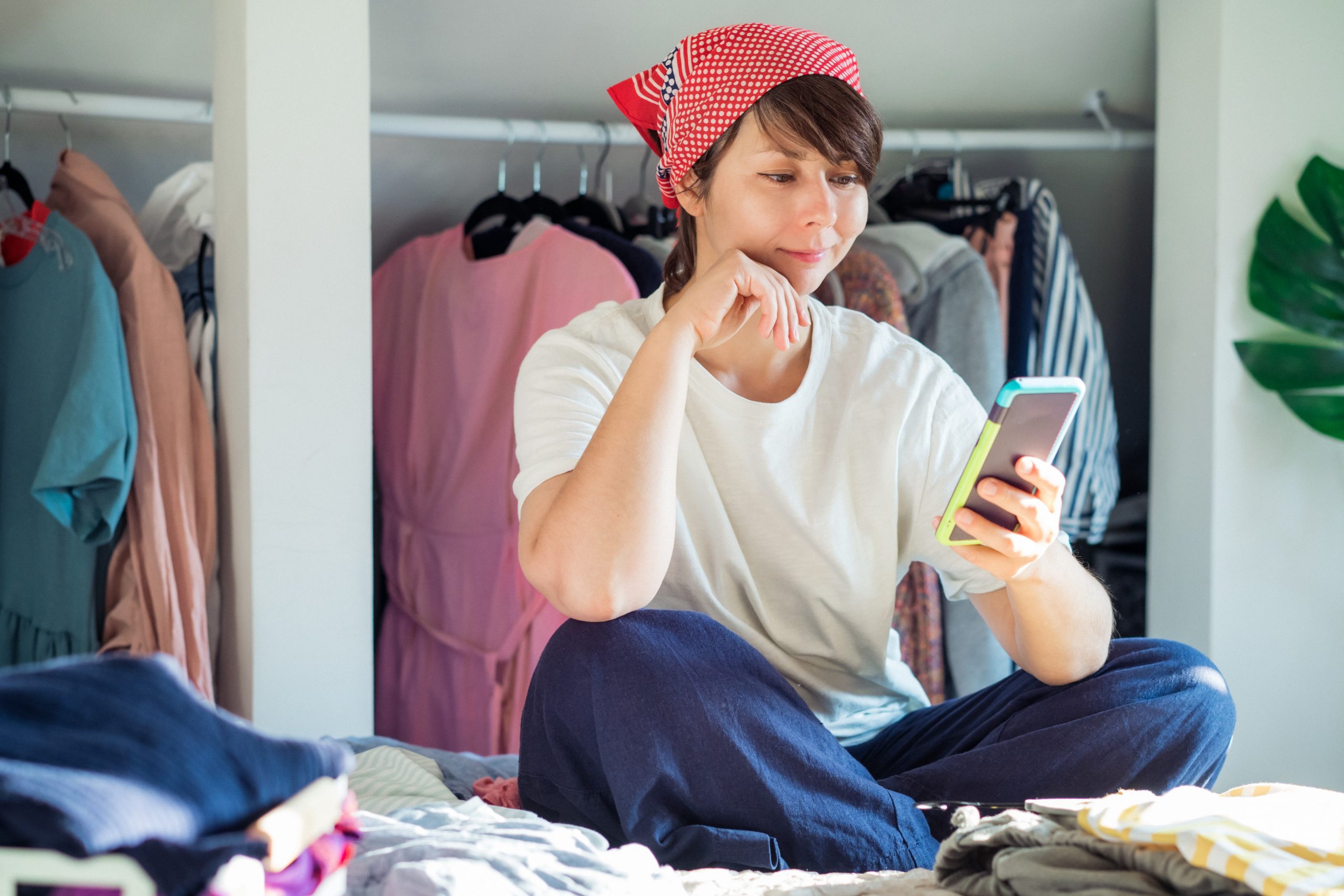 Young woman sitting on bed looking at phone, sorting through clothes
