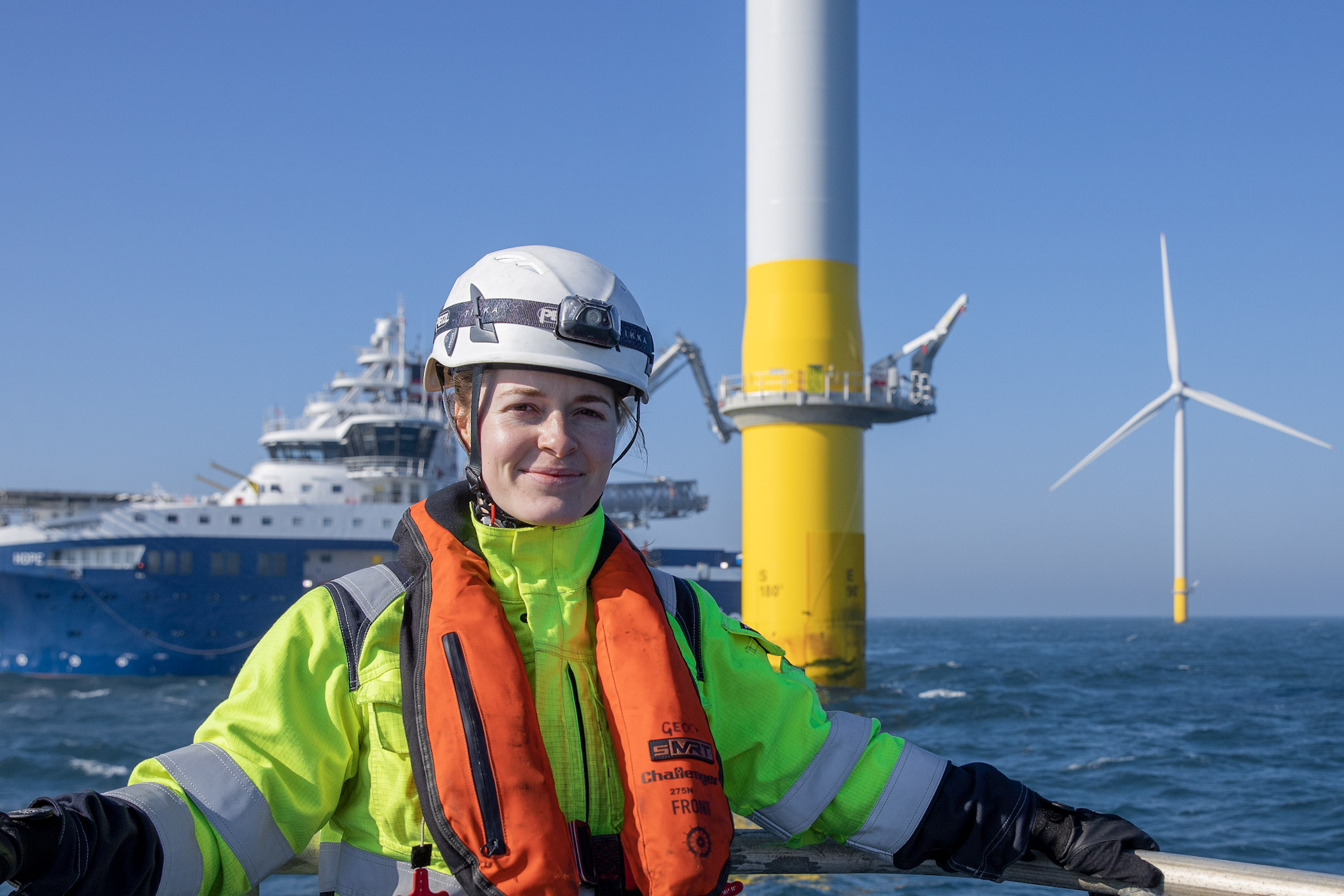 a woman in safety equipment standing on a platform in the middle of the sea in front of a ship and two wind turbines, Hornsea wind farm