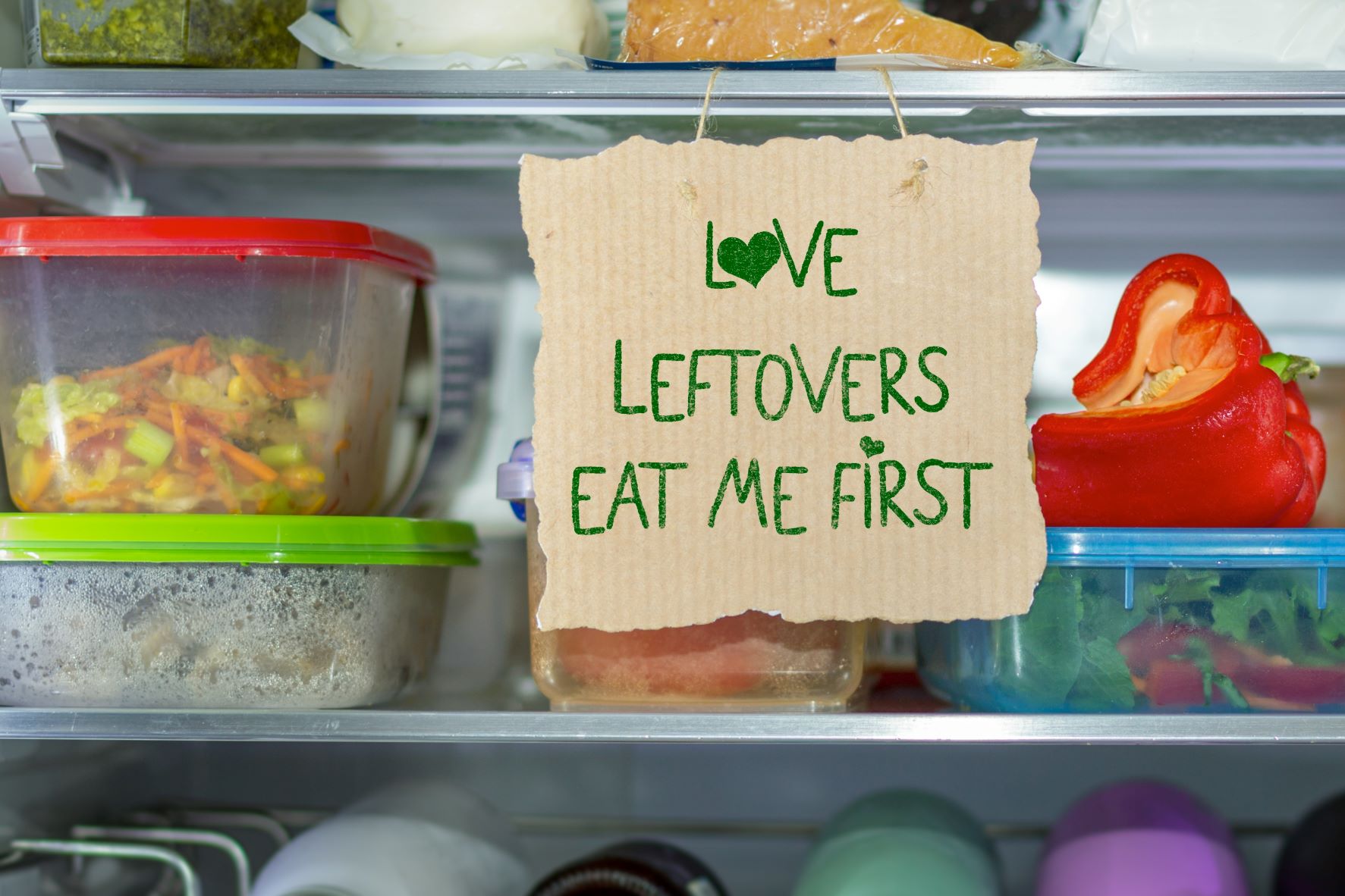 Fridge interior with 'Love Leftovers Eat Me First' label hanging from a shelf