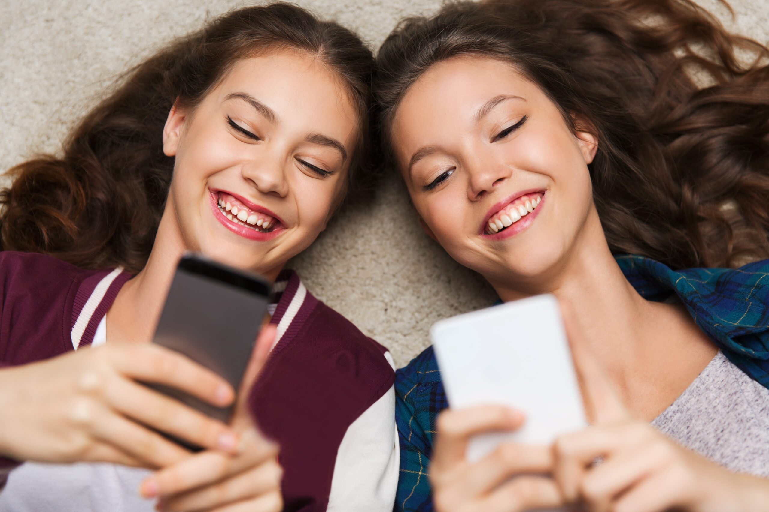 Two smiling teenage girls lying on their backs side by side looking at their smartphones
