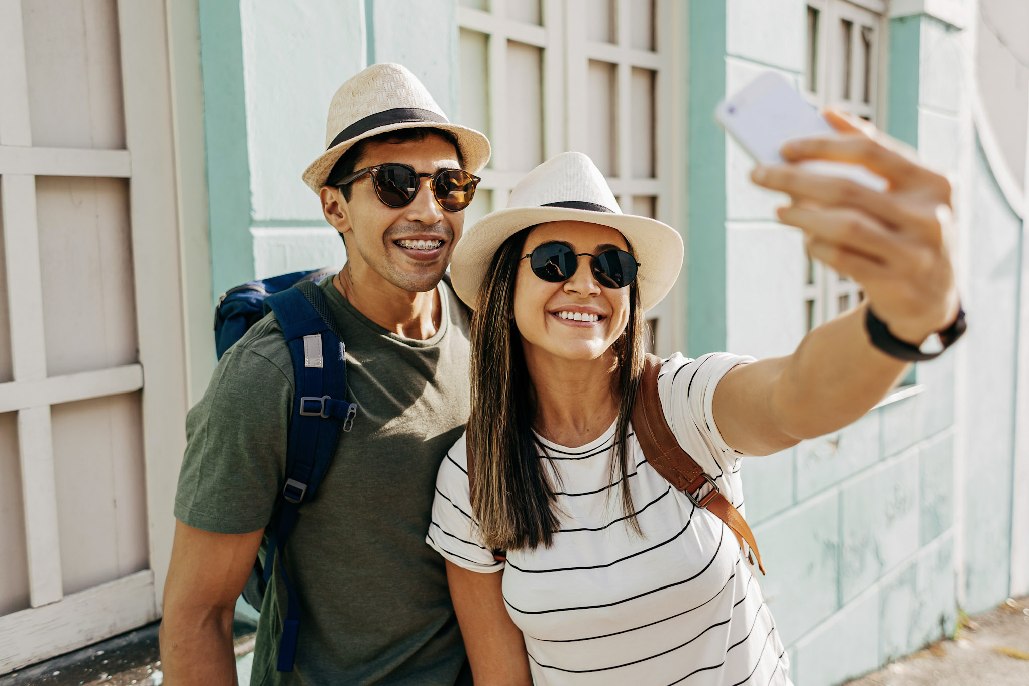 stock photo of a tourist couple in sunglasses and sunhats taking a selfie together using a smartphone while outside in a sunlit street