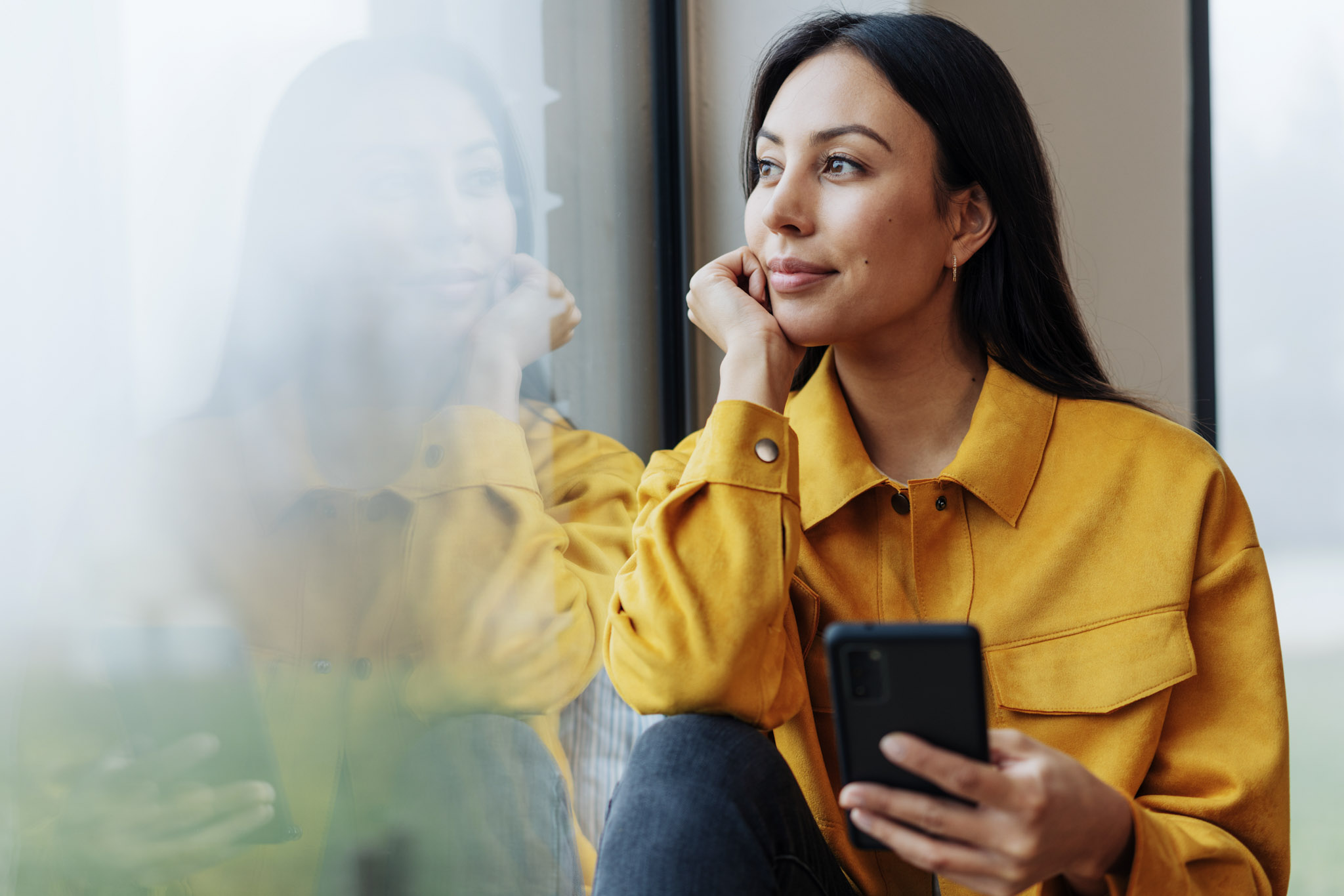 stock image of a woman looking wistfully out of a window with a smartphone in her hand