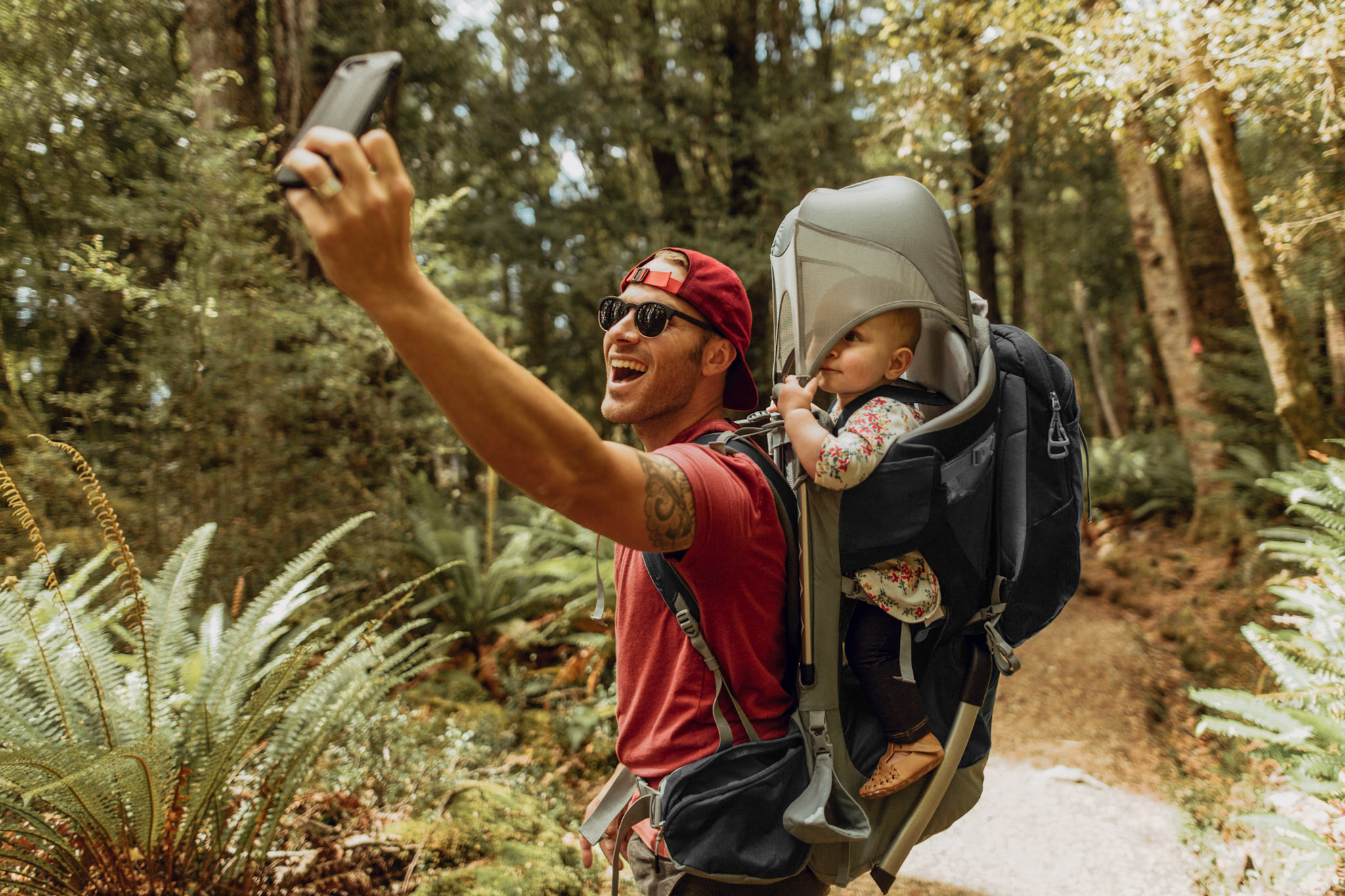 stock image of a young father hiking with their child in a carrier on their back while taking a selfie