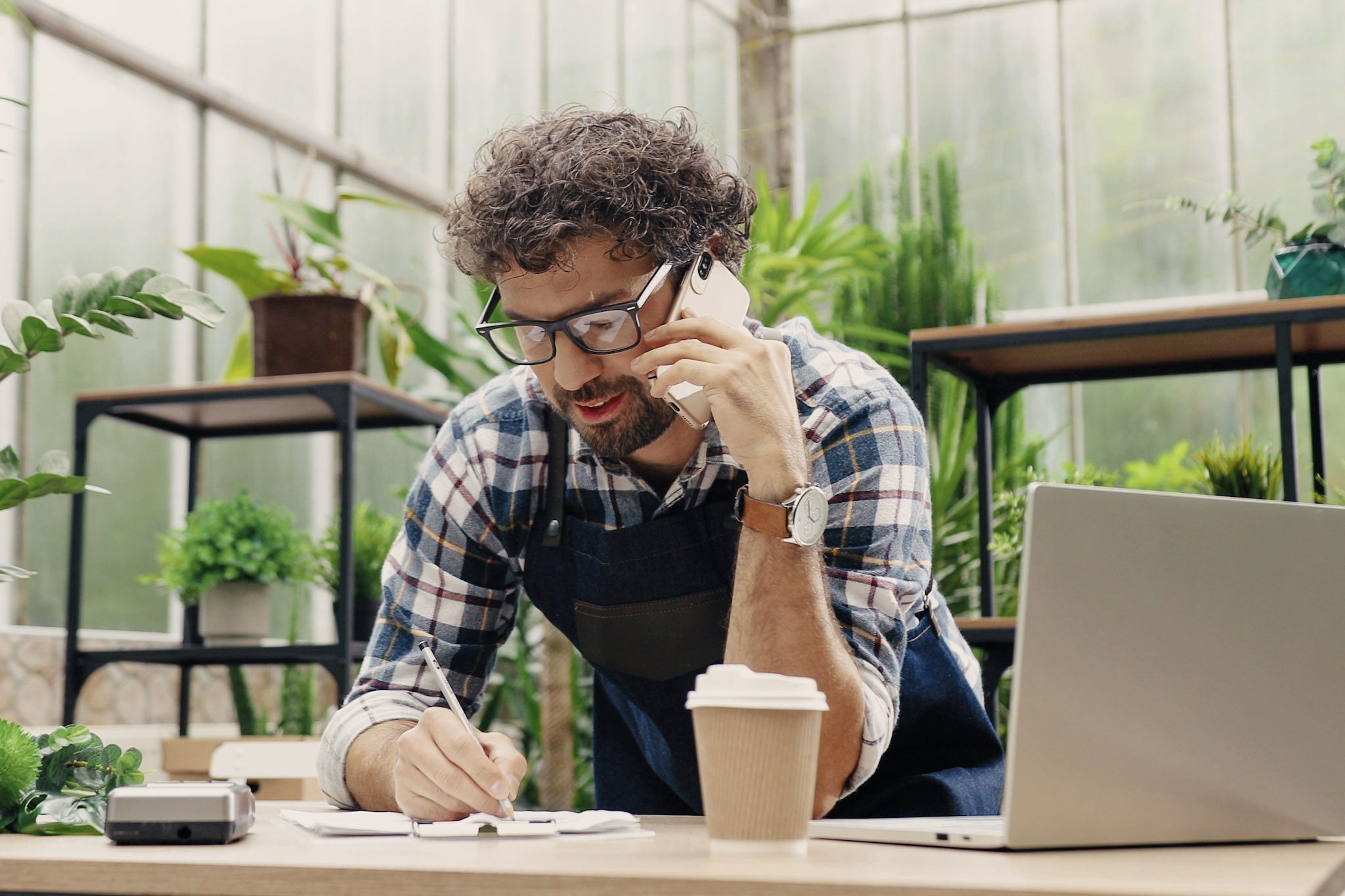 stock image of a small business owner talking on a smartphone and taking notes while surrounded by plants and standing in front of a laptop