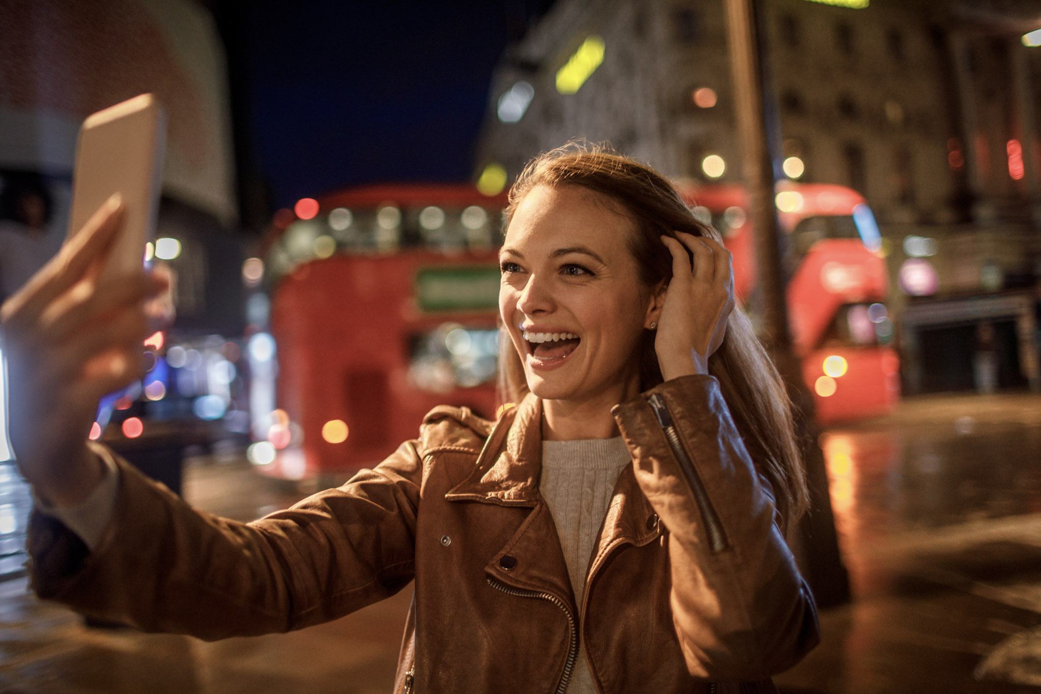 stock photo of a young woman taking a selfie at night in London with a red double decker bus passing by behind her