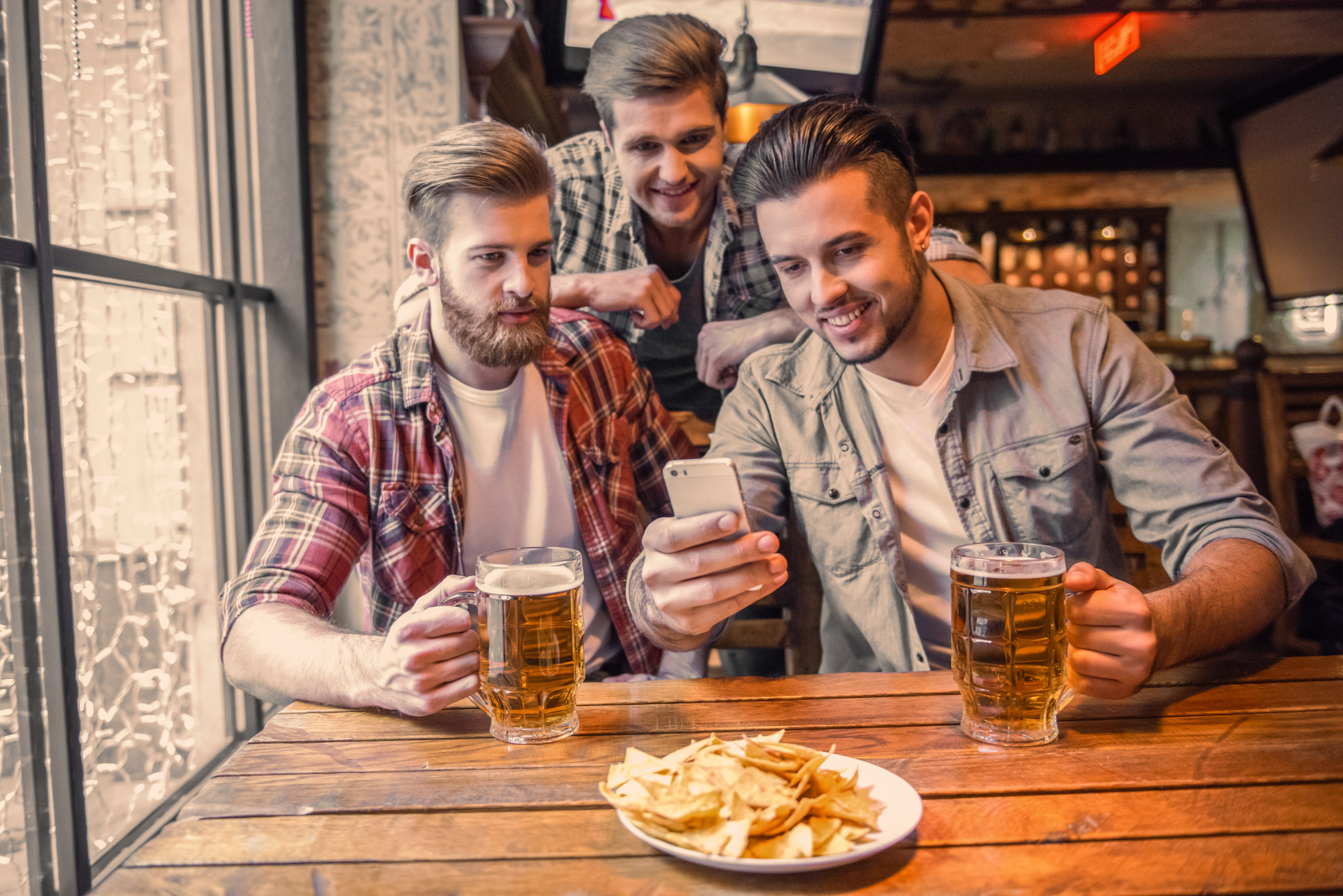 stock photo of three bearded men with excessive amounts of gel in their hair, drinking beer in a pub while looking at a smartphone