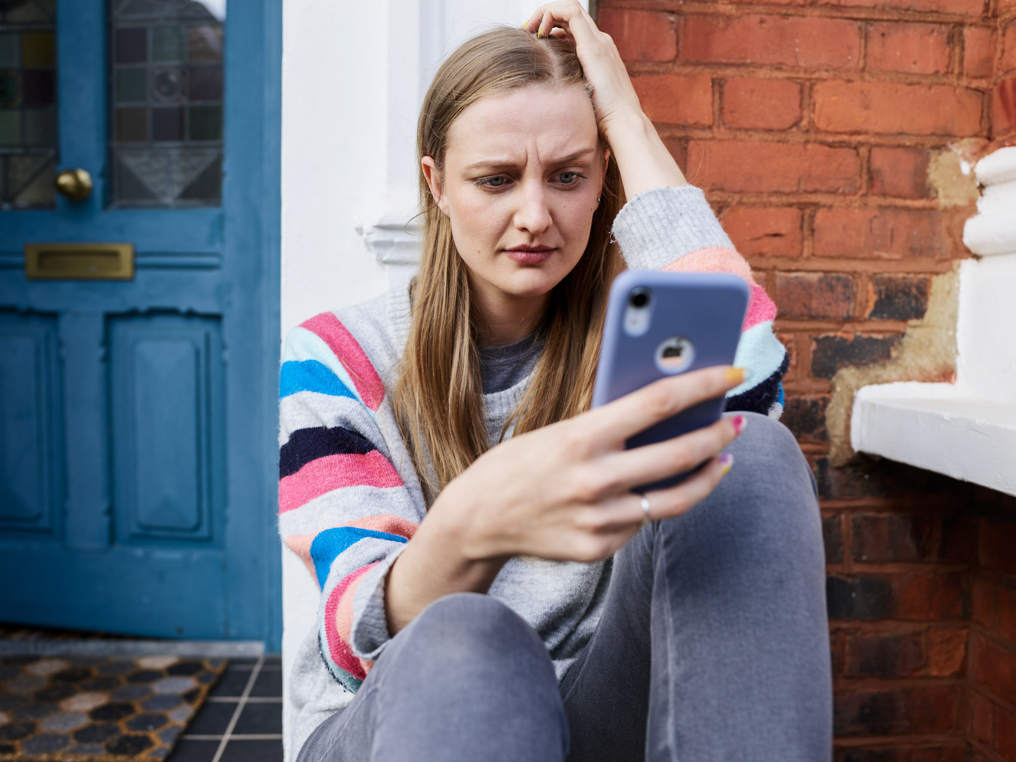 a worried-looking woman sitting on a doorstep while looking at a smartphone
