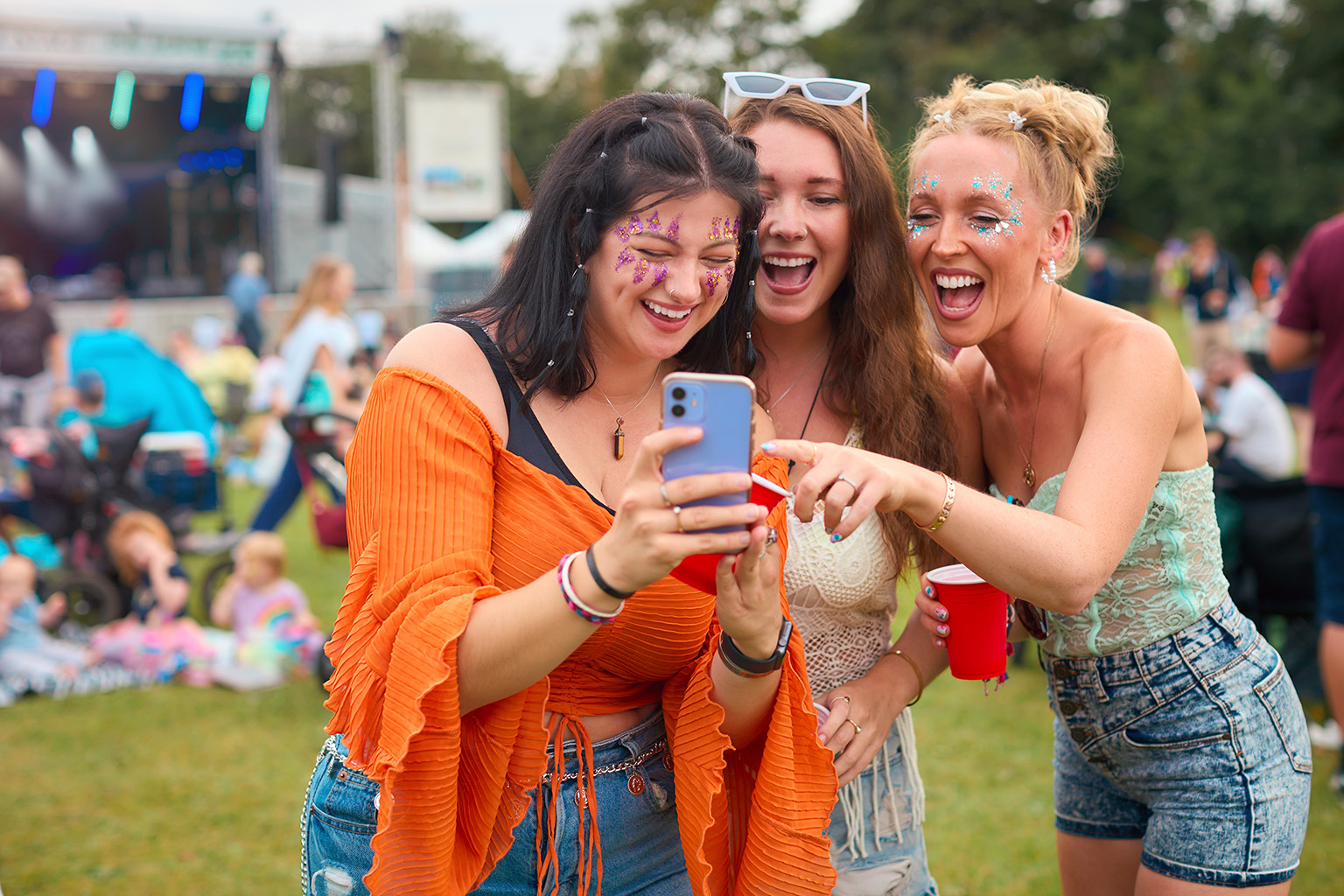 a stock photo of three women at a music festival with a stage visible in the background and one of the women holding a smartphone