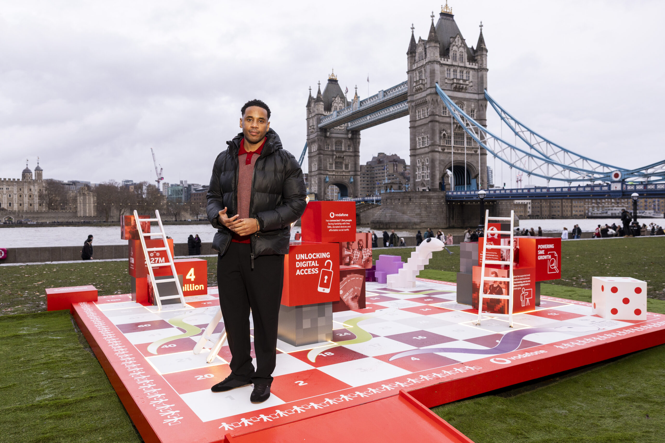 Voda-Everyone.connected-press-7 (1) Reggie Yates standing on Vodafone UK's Snakes & Ladders installation, with Tower Bridge behind him.