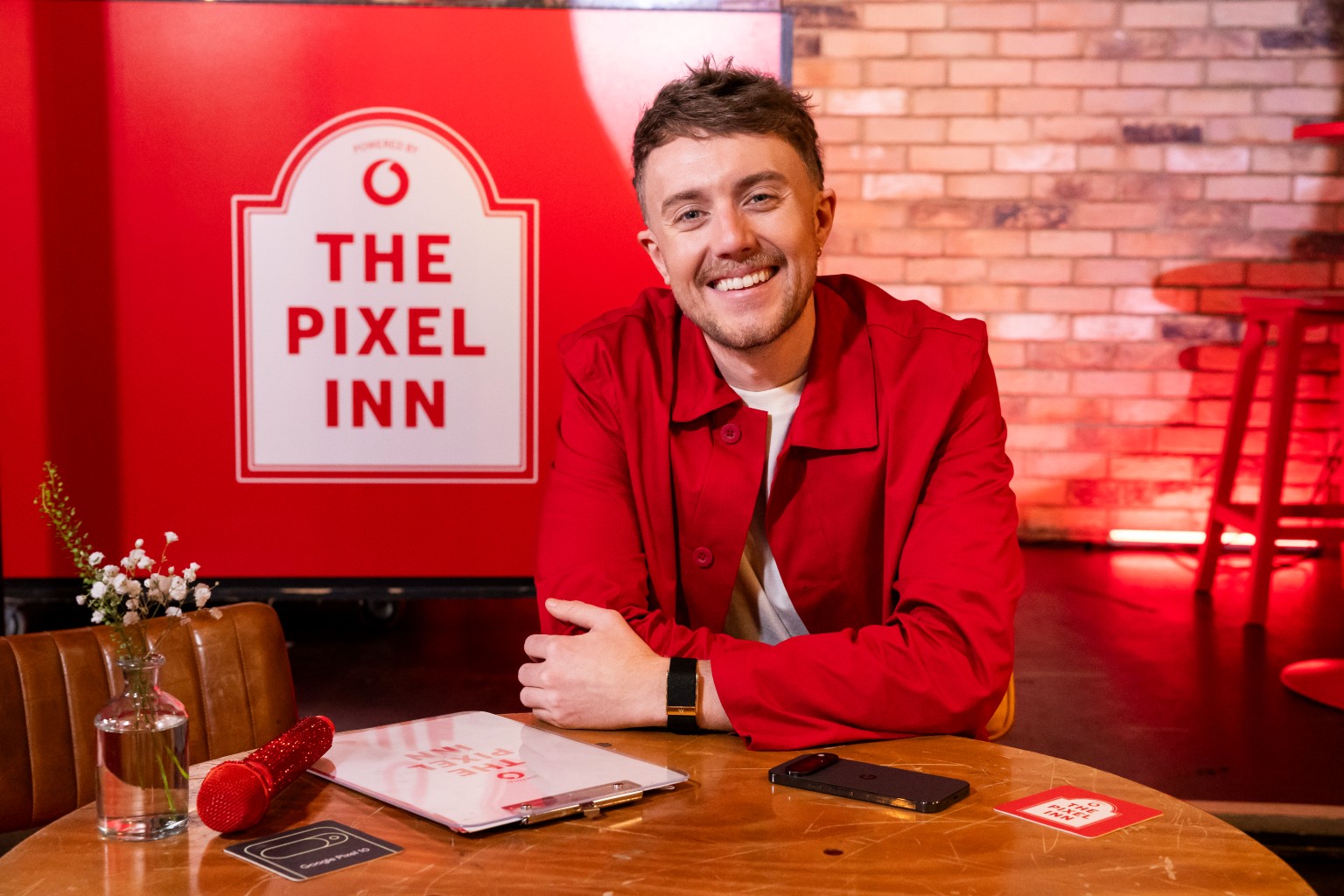 Roman Kemp sat at desk with Vodafone-branded sign behind, reading 