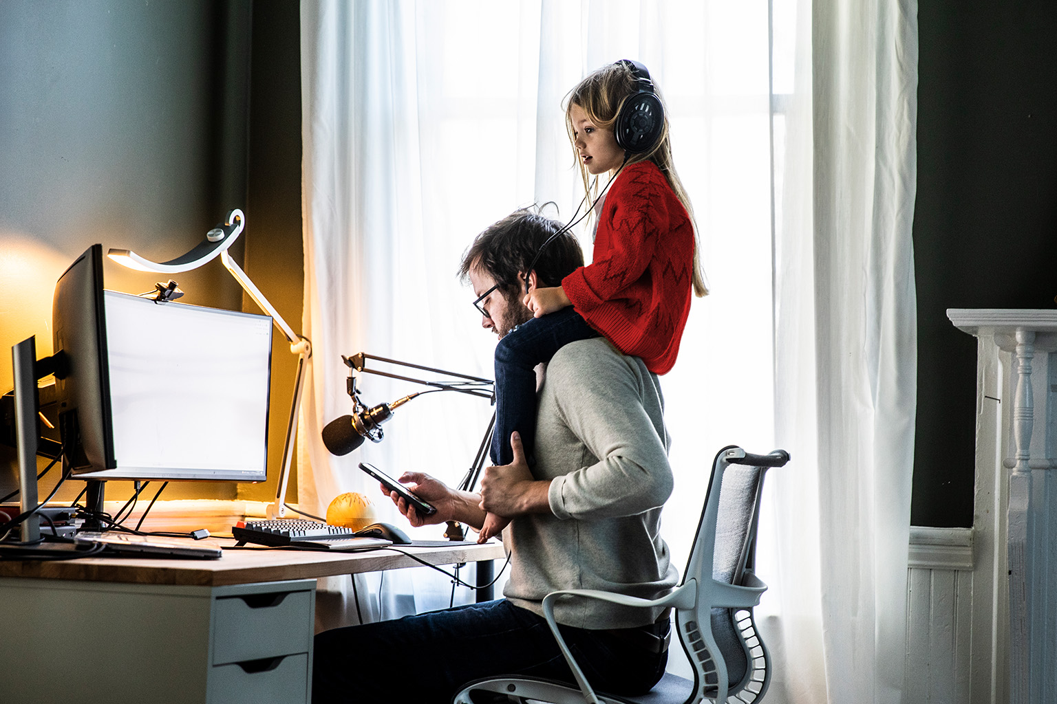 stock image of a young girl in a red sweater sitting on her father's shoulders while he sits a desk using a smartphone in front of a desktop computer on a desk
