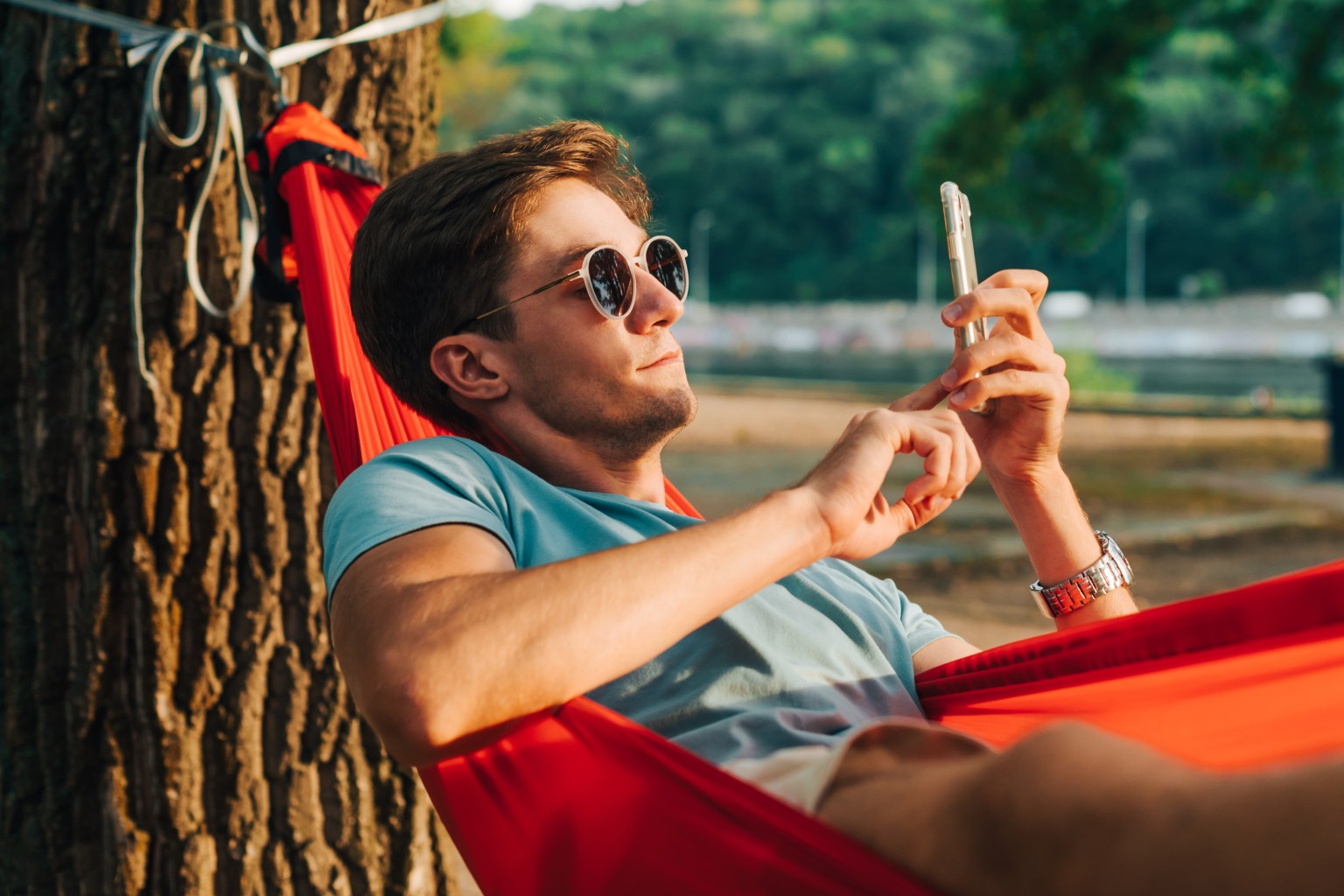 Young man in sunglasses lying at sunset in a hammock and using a smartphone.