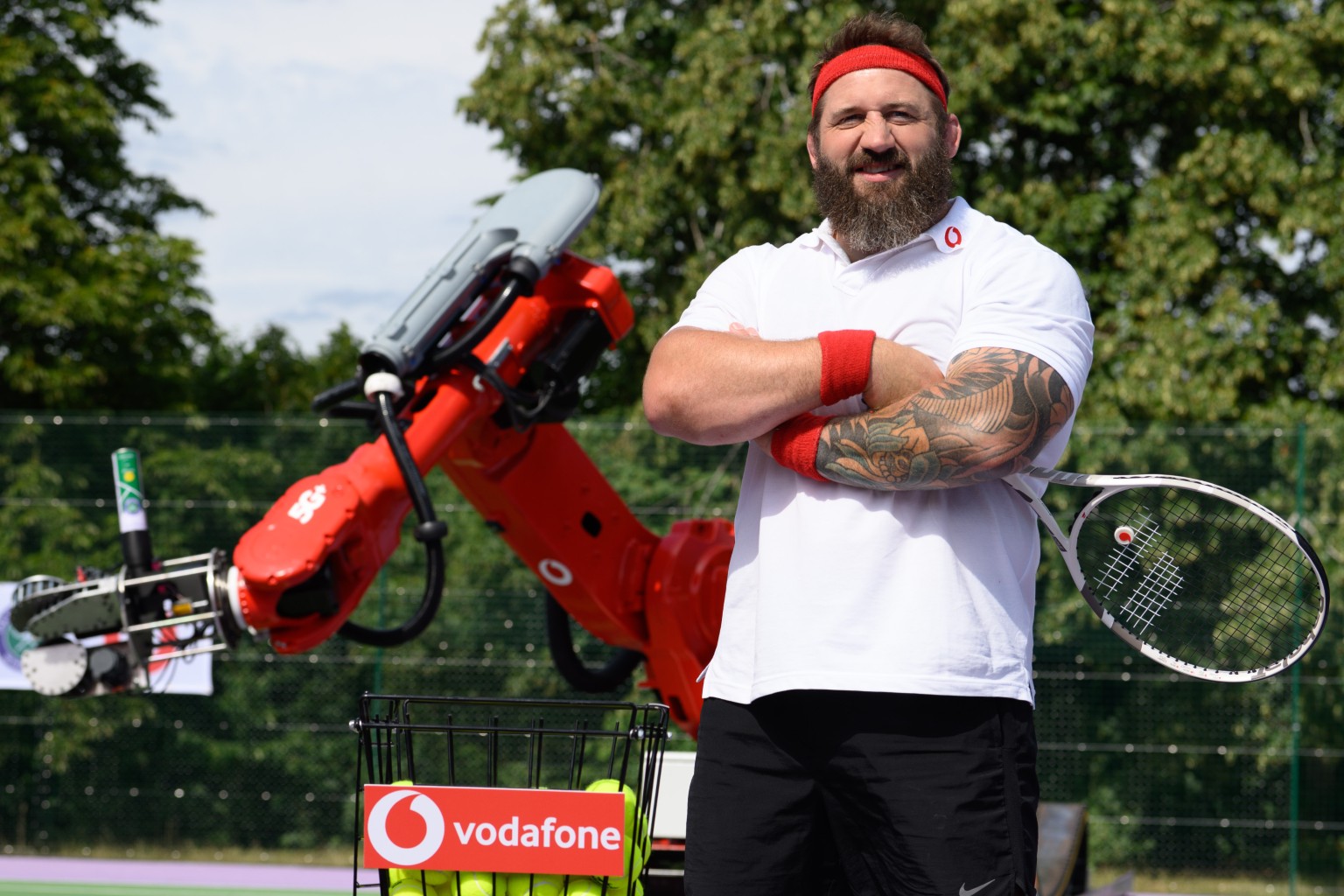 Joe Marler posing in tennis outfit, in front of Wimbledon 5G+ Serve robotic arm.