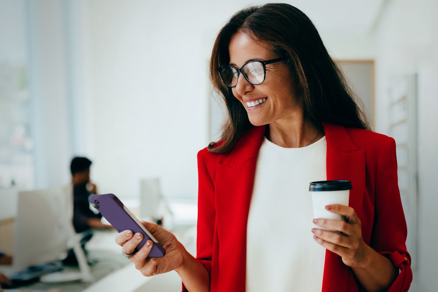 Smiling woman in red blazer holding coffee and looking at smartphone.