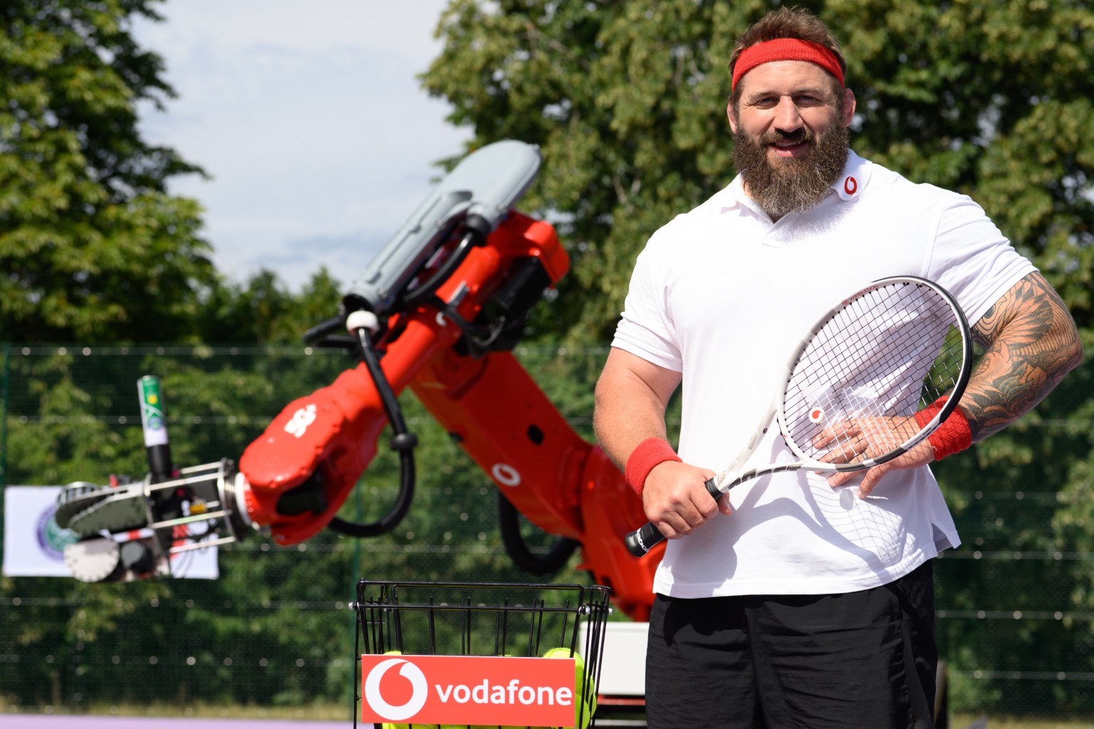 Joe Marler posing in tennis outfit, in front of Wimbledon 5G+ Serve robotic arm.