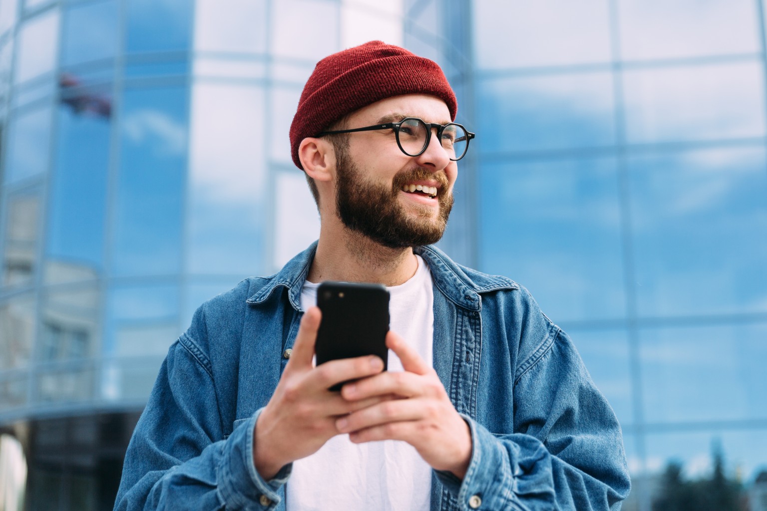 AdobeStock_361859073 (1) Man with red hat, beard and glasses smiling while using phone.