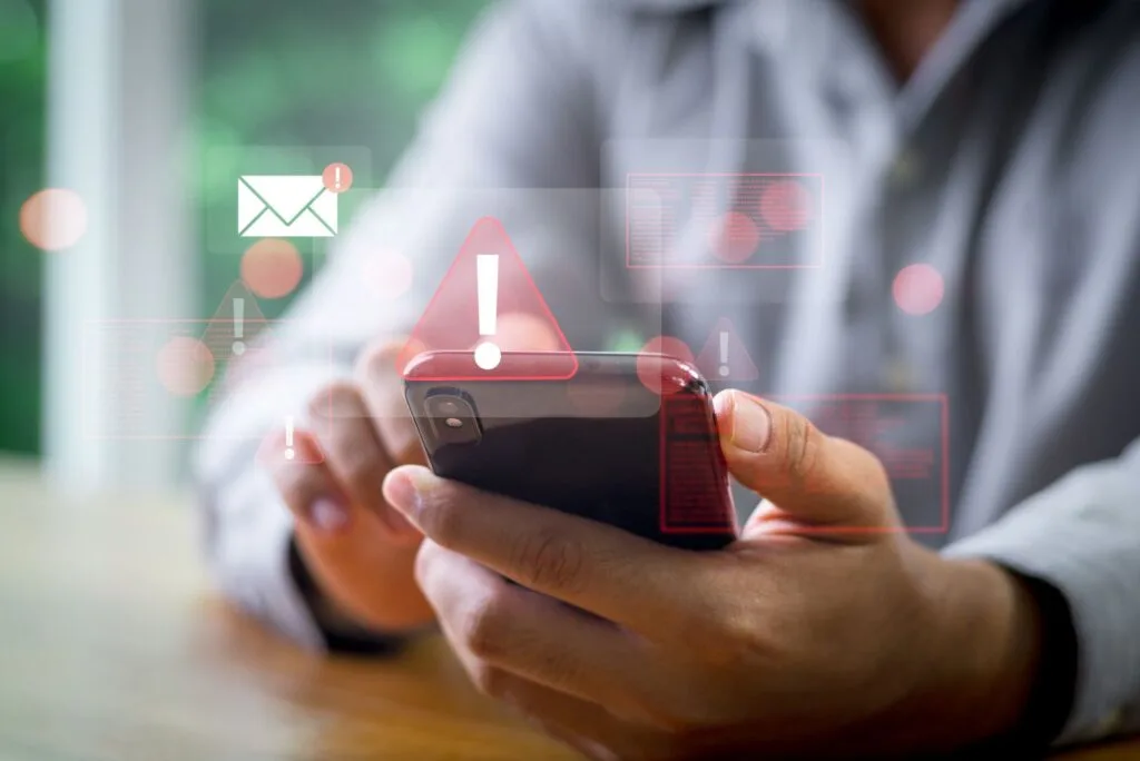 Hands holding a smartphone surrounded by digital warning signs, an email icon with an alert, and data overlays, suggesting security concerns.