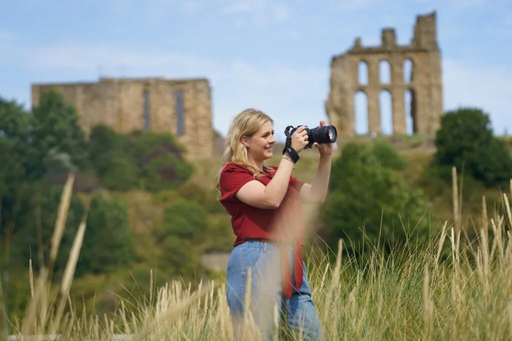 A smiling blonde woman in a red top and blue jeans holds a camera to her eye in a grassy field, with ancient stone ruins on hills in the background.