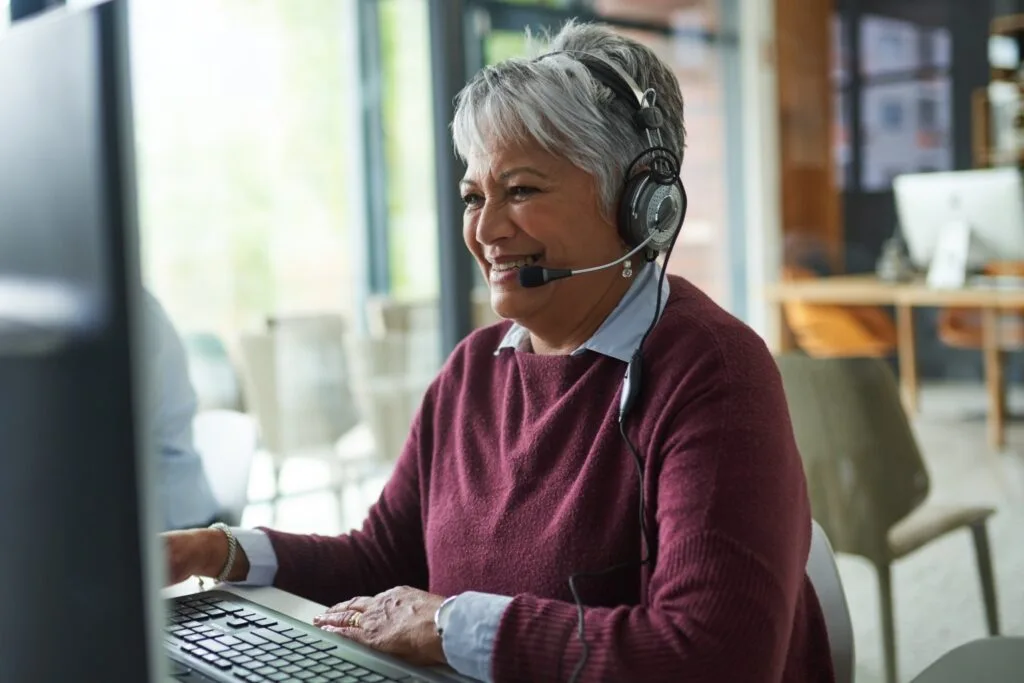 AdobeStock_495352616 An older woman with grey hair smiles while wearing a headset and typing on a computer keyboard.