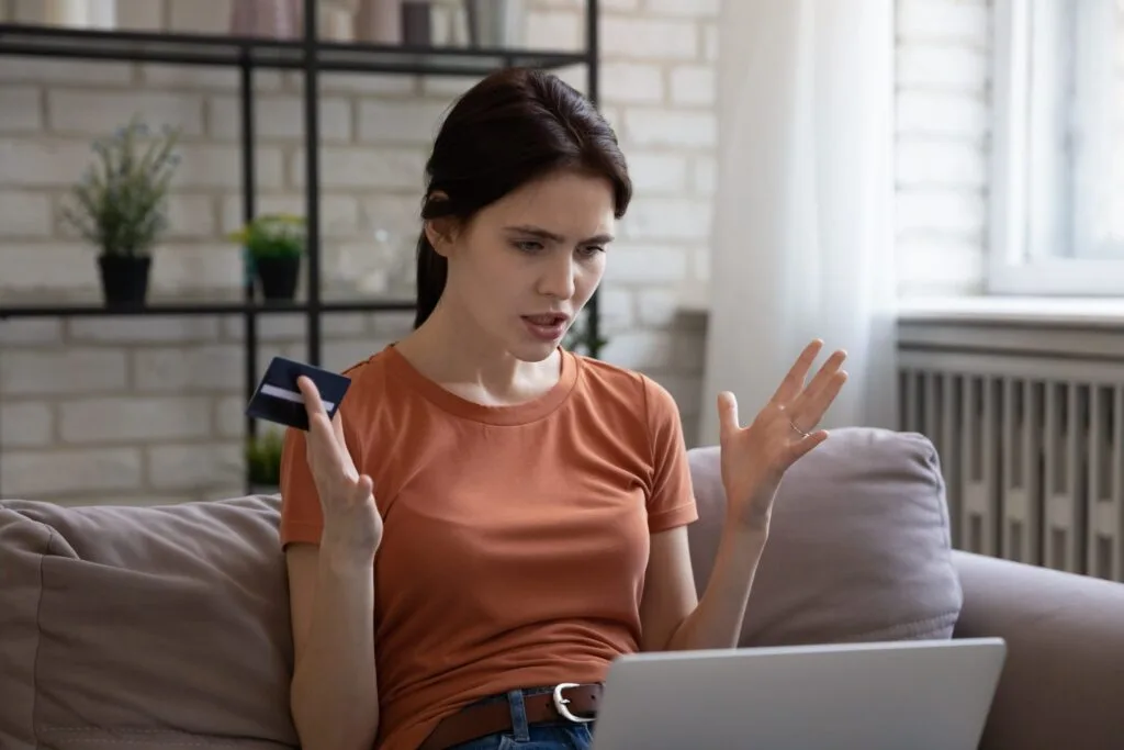 shutterstock_1800910465-scaled A frustrated young woman sits on a sofa, holding a credit card and gesturing at a laptop.